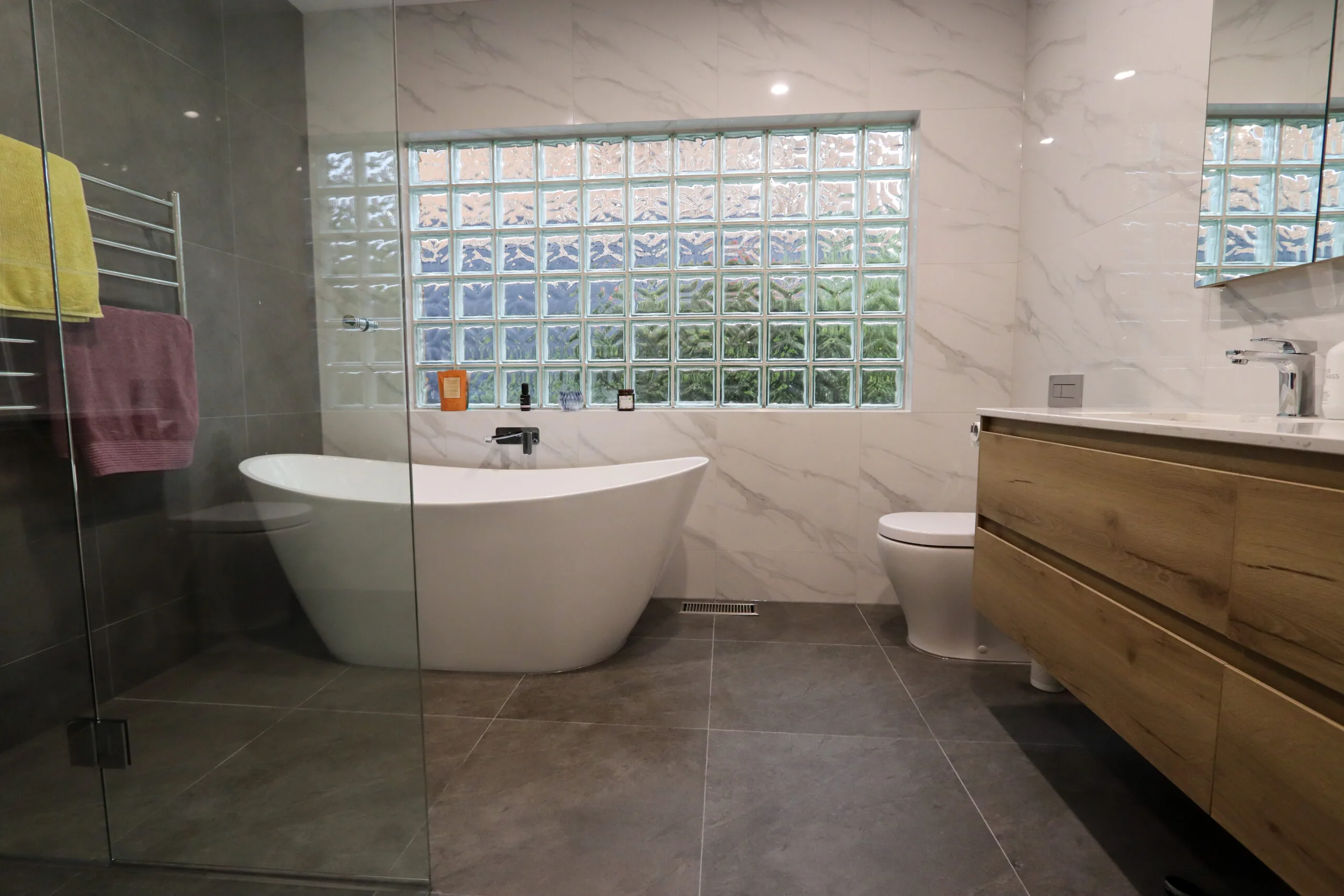 Freestanding bath beneath a glass-block window beside a timber vanity.