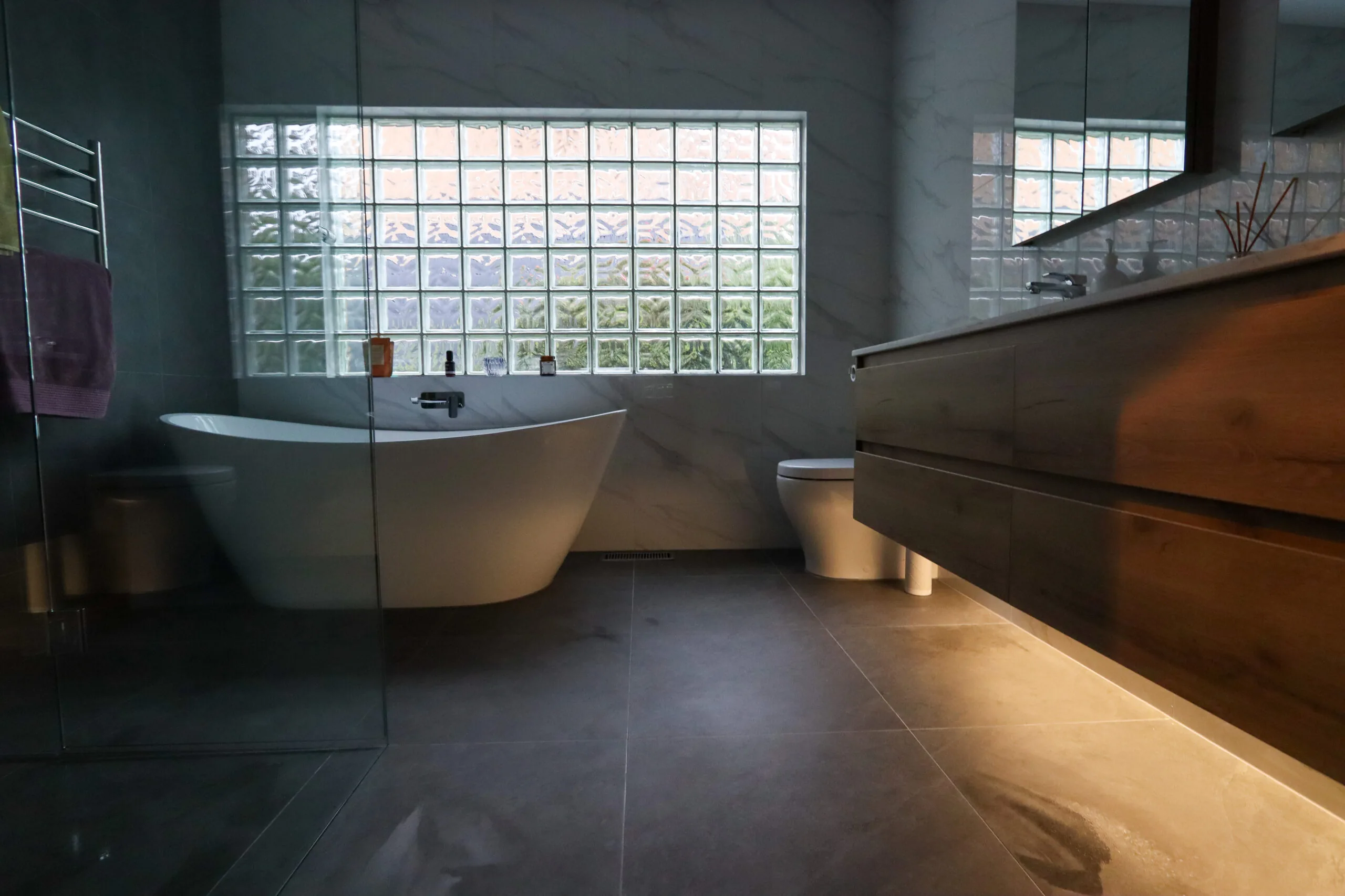 Freestanding bath beside an underlit timber vanity in a dark tiled bathroom.