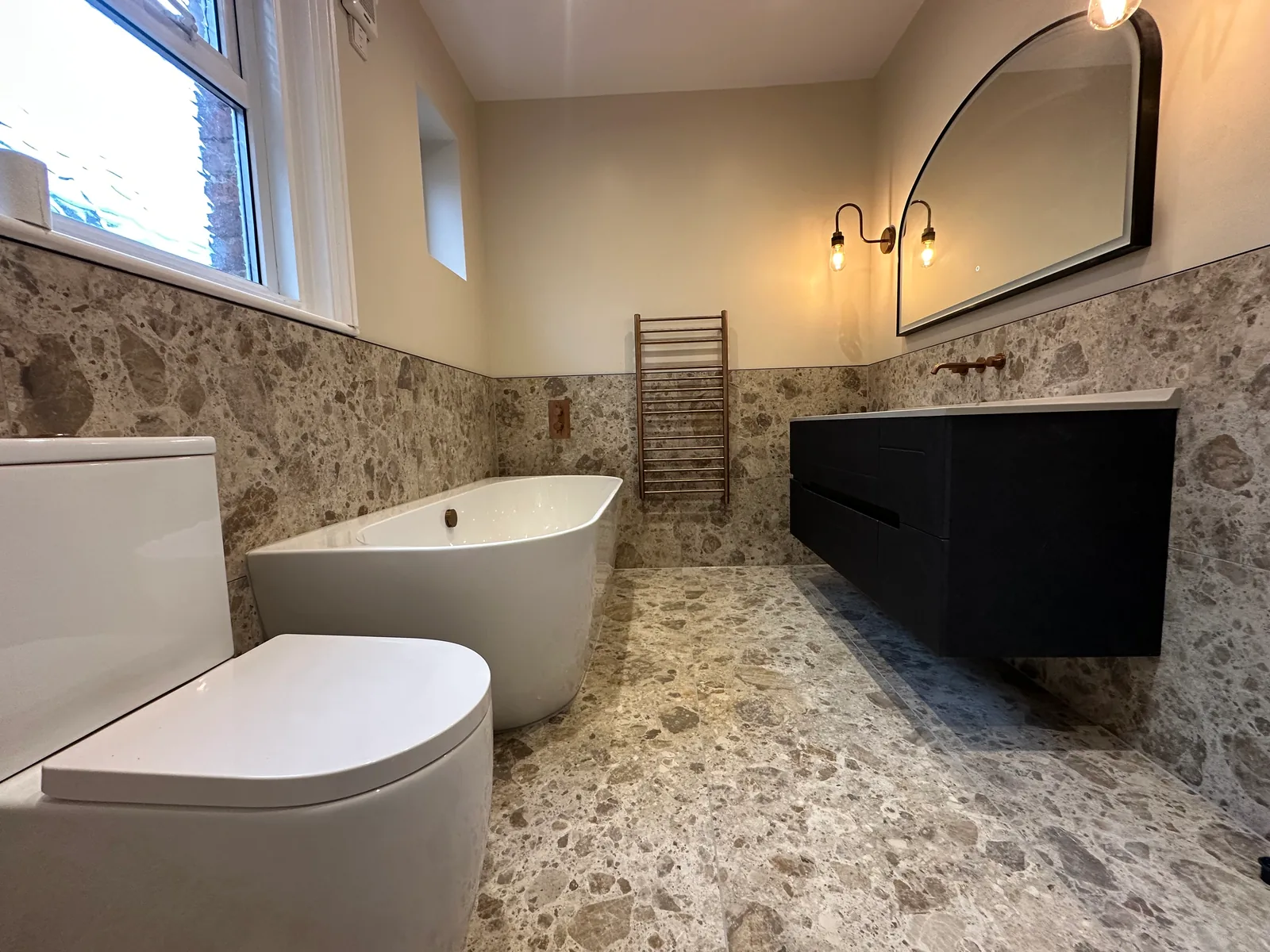 Freestanding bath and dark vanity beneath an arched mirror in the Cheltenham bathroom.